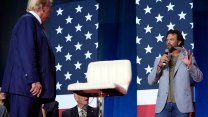 A man in t-shirt and sport jacket holds a microphone in his right hand and piece of paper between his fingers in his left hand as he stands on stage. The bearded middle-aged man is speaking in the direction of Donald Trump, who is dressed in a a navy suit and red tie. At the center of the photo is an empty stool with back rest. And, behind the man speaking on stage are panels dressed in the pattern of the American flag. Audience members on the left side of the image, look at the men.