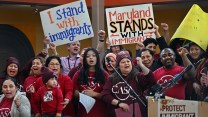 A group of people cheer. Some hold signs that read, "I stand with immigrants," and, "Maryland stands with immigrants."