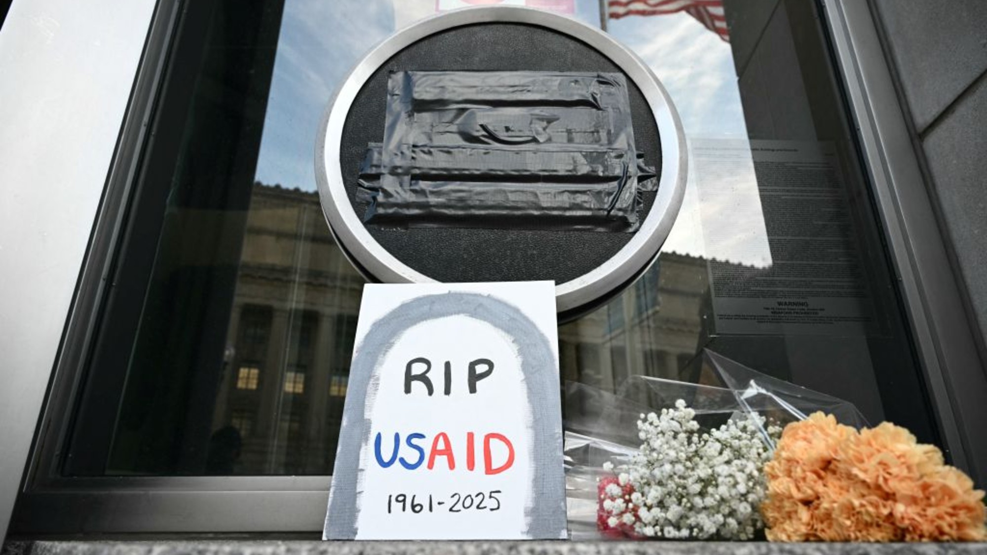 A trash bag is covering the USAID sign at the agency's DC headquarters. A poster in front says "RIP USAID."