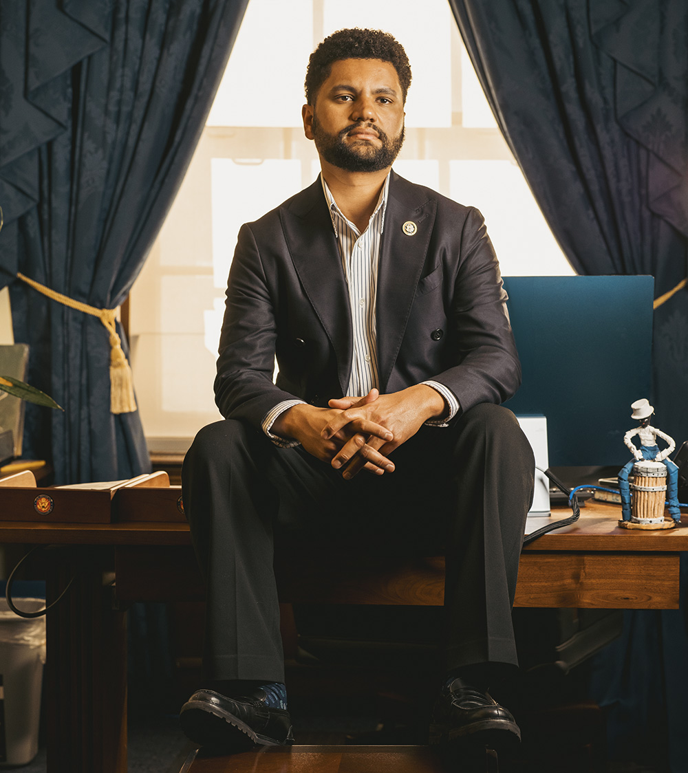Representative Maxwell Frost sits on his desk in his office in D.C.; his expression is serious and his gaze looks off beyond the viewer.