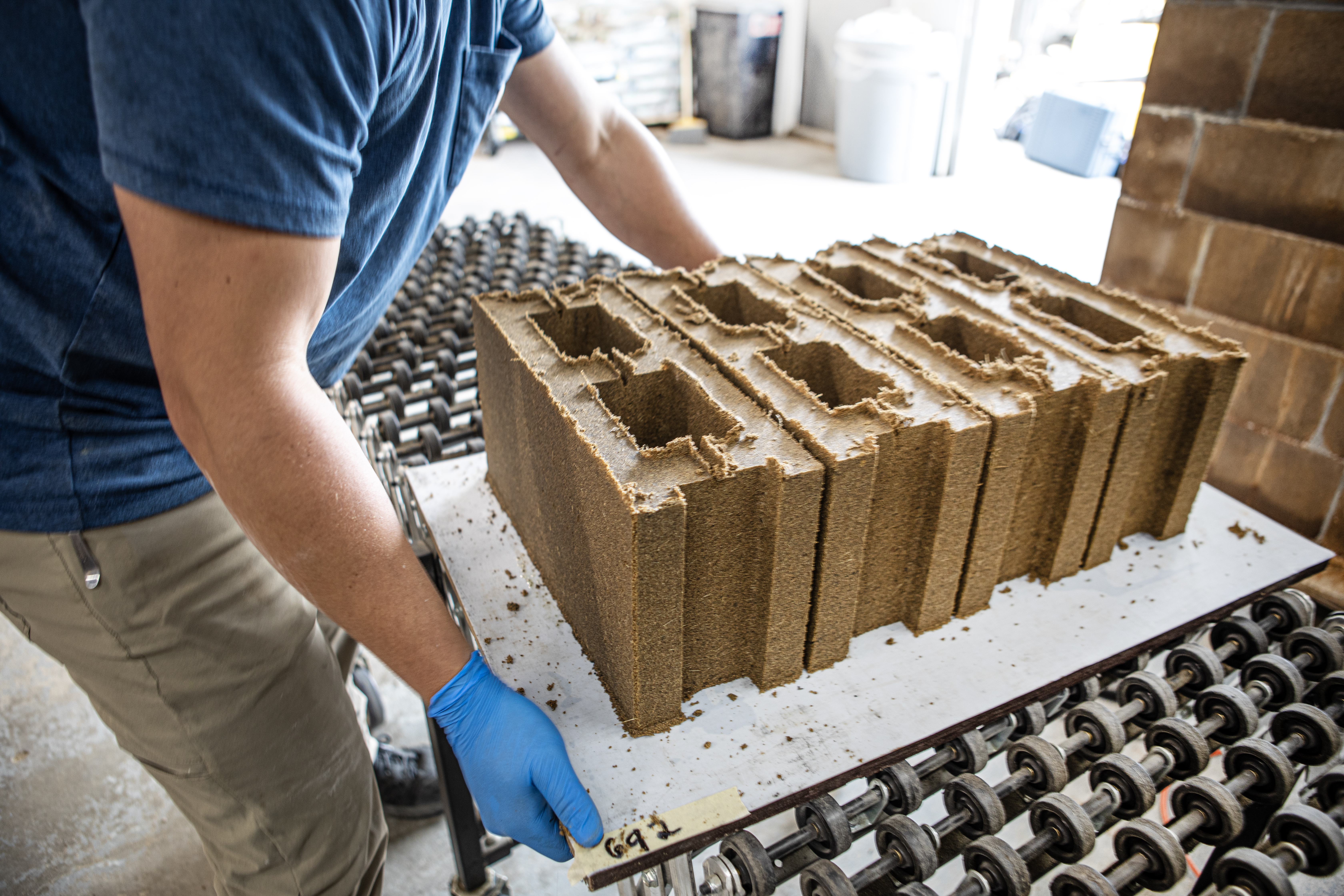 Brown concrete blocks sit on a table. A man wearing a blue shirt and blue gloves bends over them.