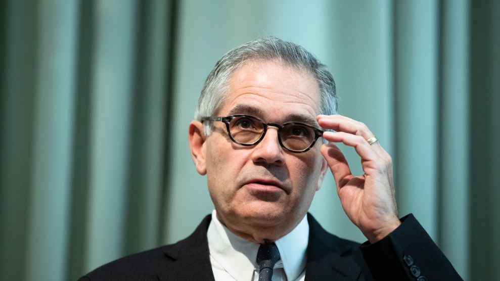 A middle-aged man with graying hair, suite and tie reaches for the frame of his glasses with his left hand. His head is tilted slightly downward, and he has a serious expression on his face. Behind him is a green curtain backdrop.