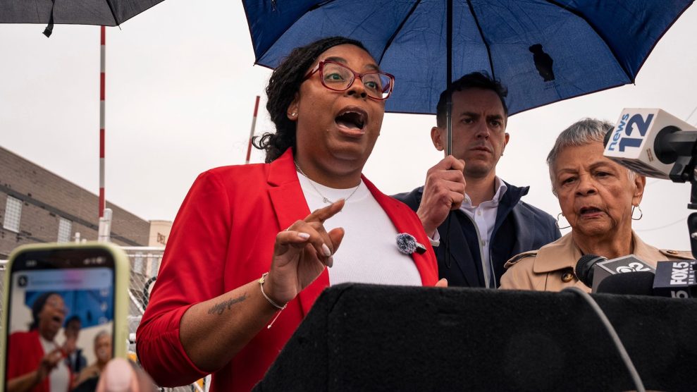Three people standa at a press conference podium under an umbrella.