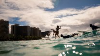 Six surfers ride the waves at Kuhio Beach in Oahu, Hawaii. Clear blue water approaches the camera lens. In the background, there is a beach shore, trees, blue sky with white clouds, and multiple buildings.