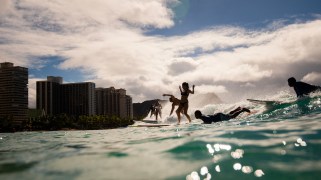 Six surfers ride the waves at Kuhio Beach in Oahu, Hawaii. Clear blue water approaches the camera lens. In the background, there is a beach shore, trees, blue sky with white clouds, and multiple buildings.