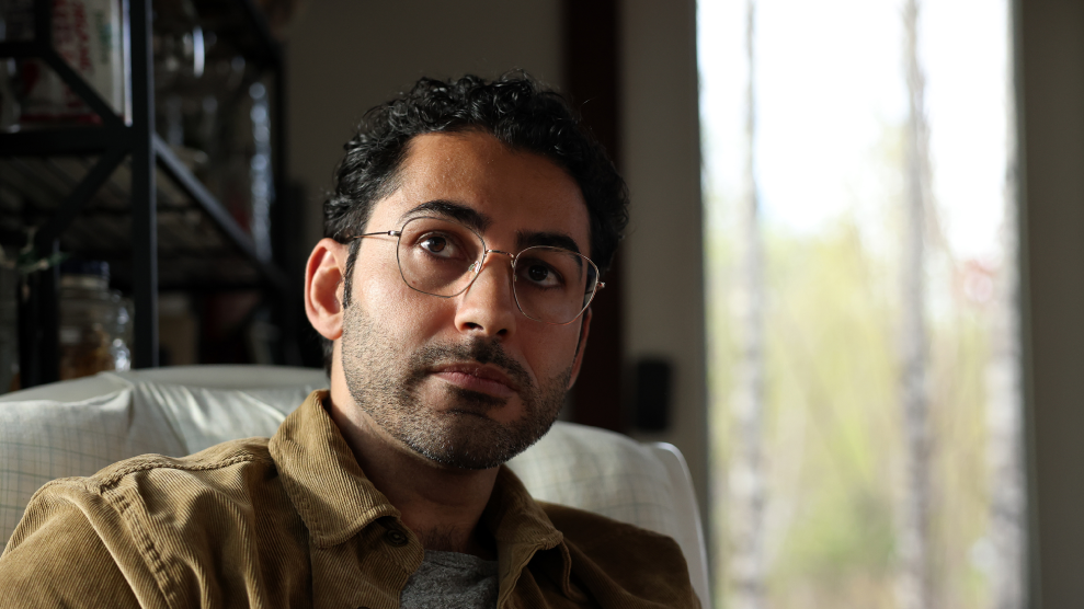 A portrait of Mohsen Mahdawi from the shoulders up. He has short, curly black hair and thin-rimmed glasses. Seated in a plush chair, he looks up at an angle with a pensive expression.