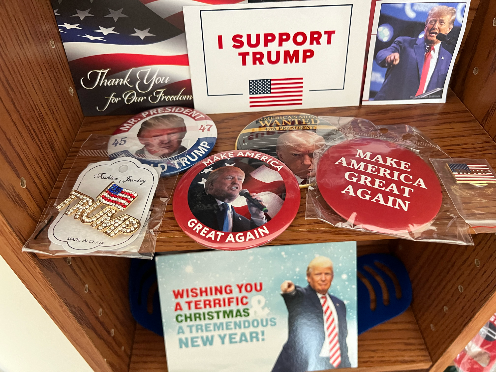 Several shelves full of tchotchkes that are branded in support of Donald Trump. There's a gold lapel pin that spells out TRUMP and above it sits an American flag.