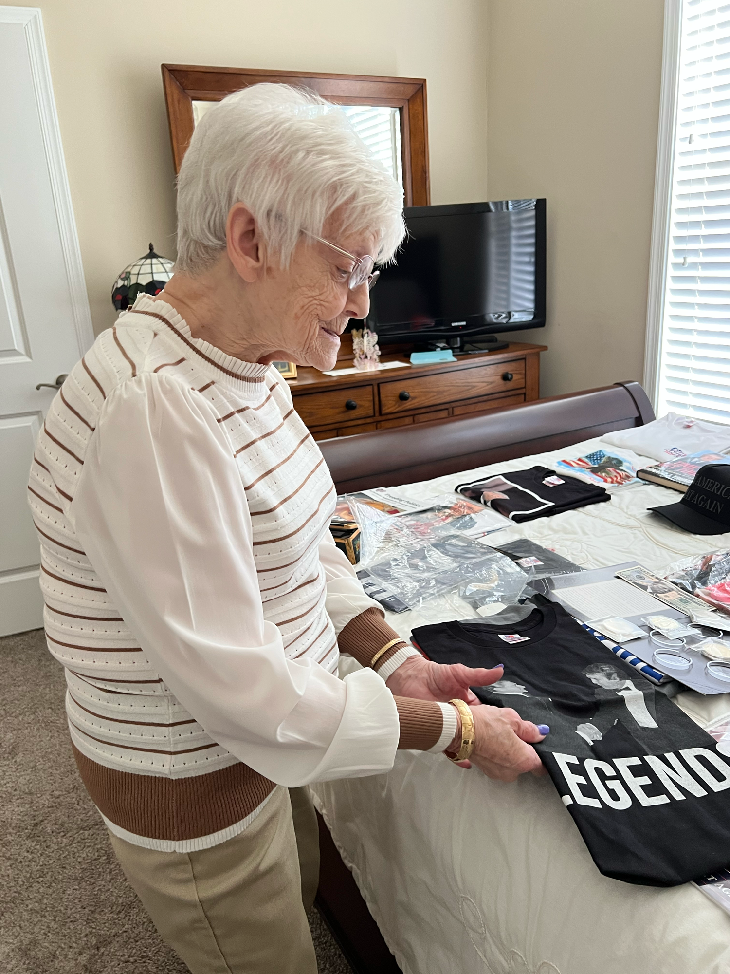 An elderly woman neatly dressed in a white blouse and tan slacks with short white hair. She stands next to a bed as she runs her fingers along a black t-shirt.