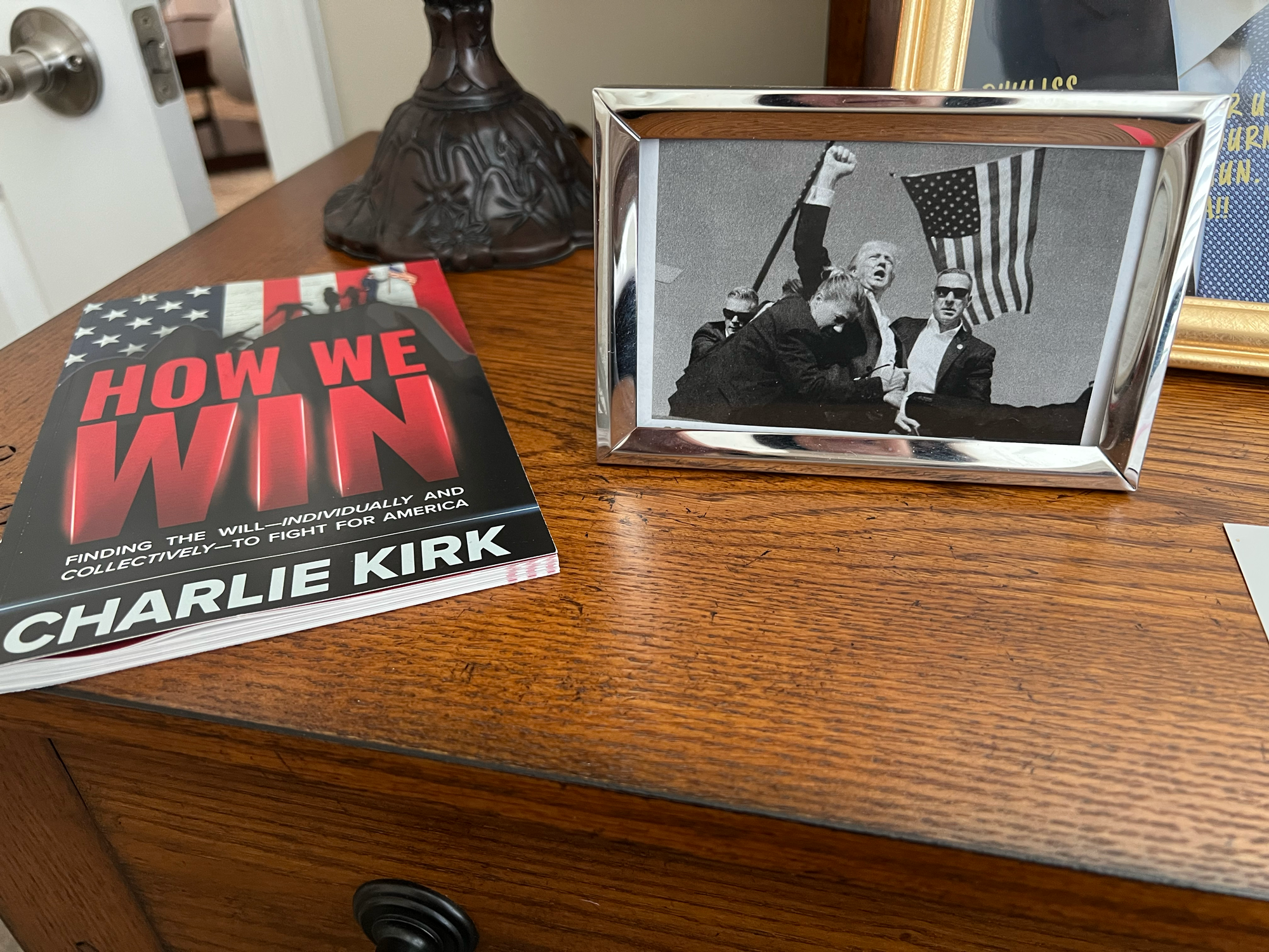 A black-and-white version of the famous photo of Donald Trump with fist raised in air as he's bracketed by secret service agents sits in a silver frame on a credenza. Beside the photo is the book "How We Win" by Charlie Kirk.
