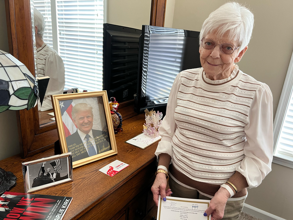 An elderly woman neatly dressed in a white blouse and tan slacks with short white hair. She stares into the camera with a smile, as she stands next to a signed portrait of Donald Trump.
