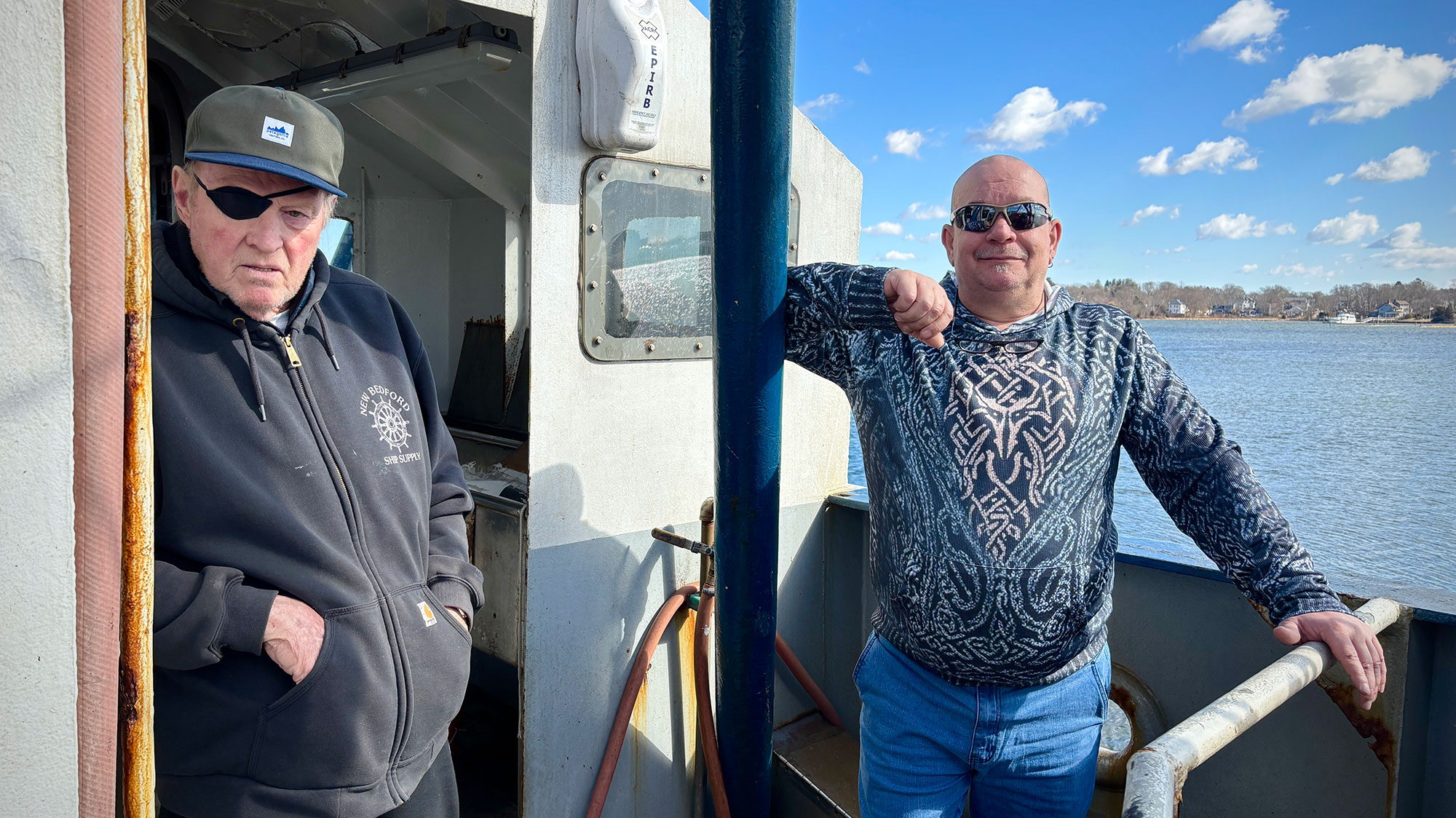 A scowling older man with an eyepatch and cap, and wearing a New Bedford hoodie, stands in the doorway of a boat's cabin, while a portly fellow in a blue sweater, with a shaved head and flavr-saver facial hair, stands outside in the sun. The portly fellow is is smiling goofily.