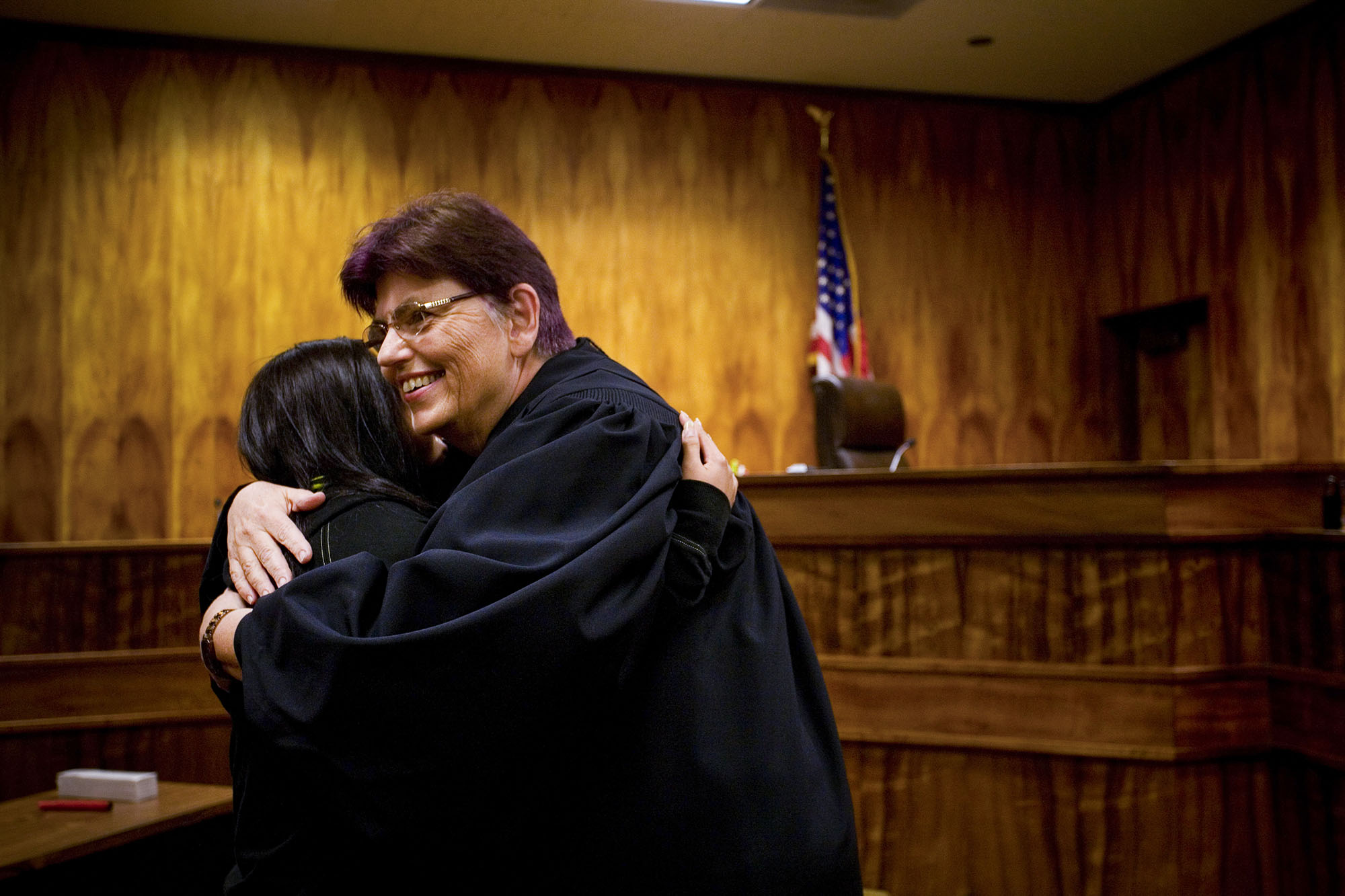 Judge Radius hugging young girl in an otherwise empty courtroom with wood paneled walls and an American flag on a pole in the background . 