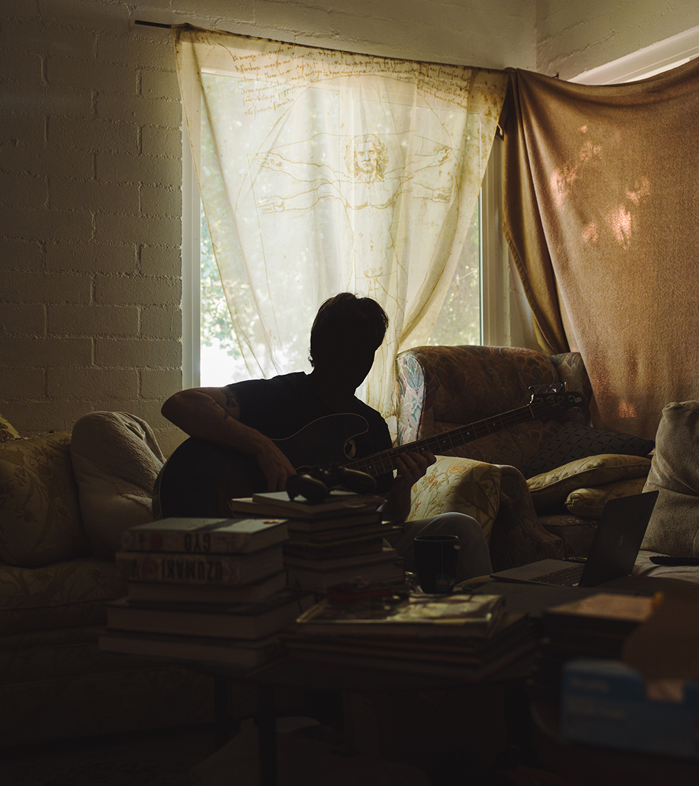 A backlit man sits on a couch near a window in a dimly lit room, holding a guitar.