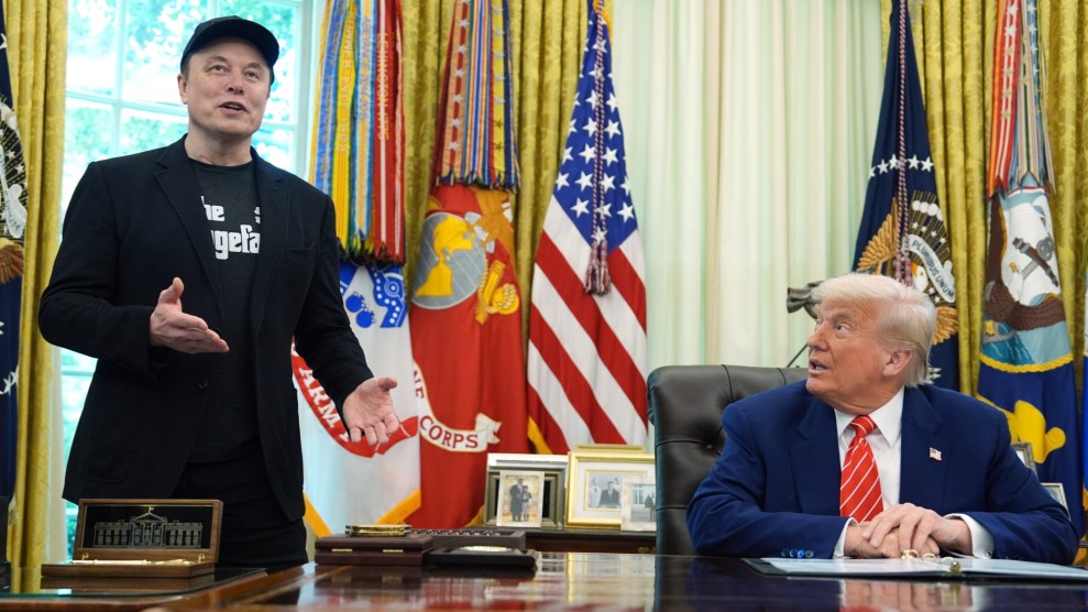 Elon Musk standing, wearing a black jacket over a graphic t-shirt and a baseball cap, next to a seated Donald Trump, wearing a navy suit and red striped tie, in front of a row of flags in the Oval Office.