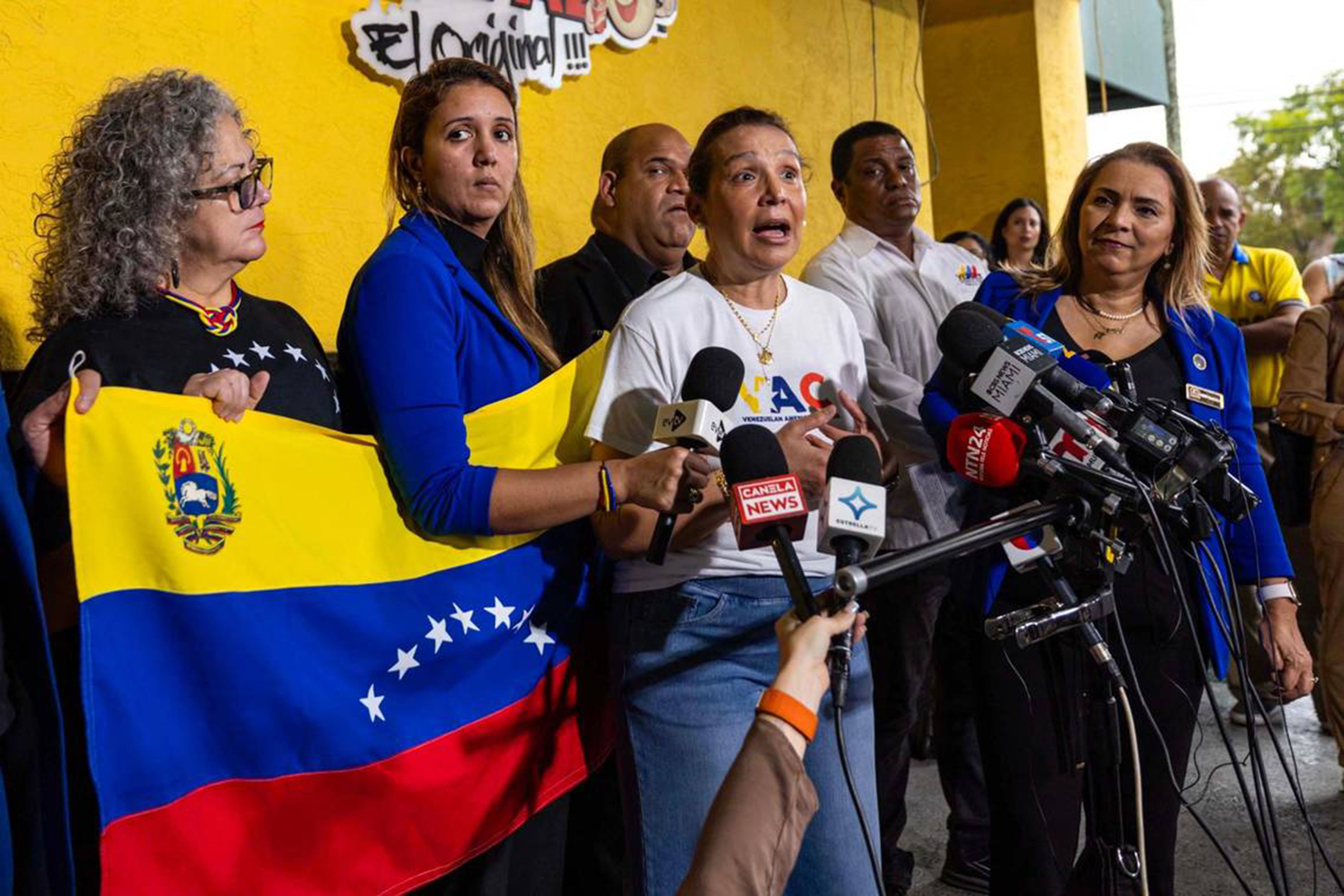 Woman speaking at a bank of microphones, surrounded by a group of people, one of them holding a Venezuelan flag.