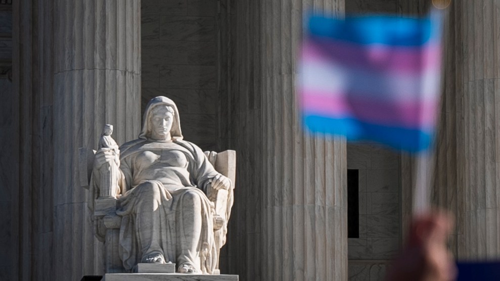 A trans flag waves in front of the statue of a seated woman at the Supreme Court.