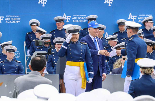 A smiling female cadet in blue dress uniform, white hat, white pants, yellow sash, salutes on a stage at her graduation from the Air Force Academy, as president Joe Biden shakes someone's hand to one side.