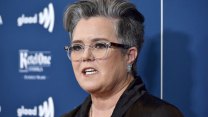 A tight shot of Rosie O'Donnell, short salt and pepper hair, clear-frame glasses, against a blue background, speaking at the GLADD Media Awards