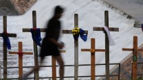A blurred figure walks past a row of wooden crosses placed along a railing in front of a rushing dam spillway. The crosses are adorned with colorful flowers, blue and white fabric, and other memorial decorations, indicating a tribute or memorial site. The scene conveys a somber and reflective mood.