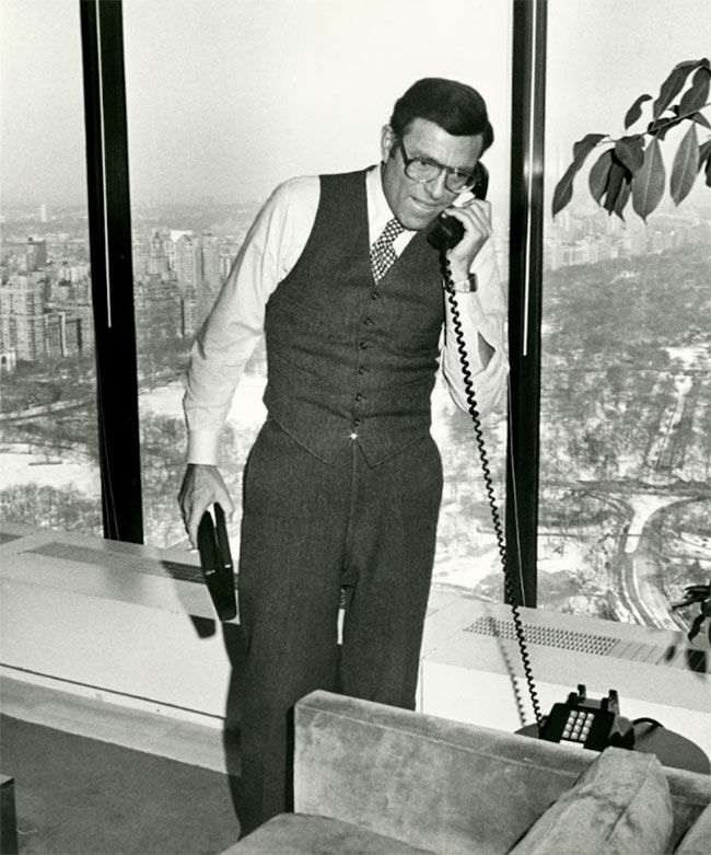 Sony executive Harvey Schein speaks on the telephone in this black and white photo. A well-groomed, handsome 1970s executive, he is standing in front of large windows looking down on the city, and is wearing a vest and checkered tie.