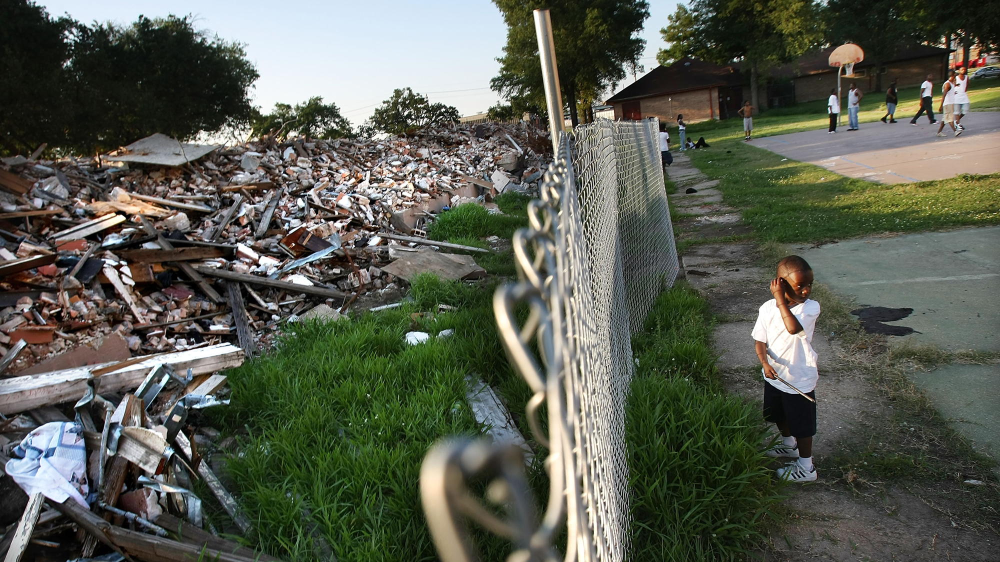 A photo divided by a chainlink fence. On the right we see people playing basketball on a blacktop in the background. In the foreground is a child in a white t-shirt and shorts looking back over his shoulder. On the other side of the photo, and the fence, is a massive pile of debris.