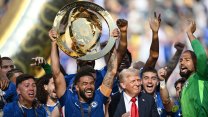 Smiling soccer players in blue jerseys on a sunny day hold a golden club world cup trophy aloft as Donald Trump embarrasses himself by crashing their party