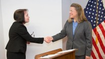 Annie Caputo and Kristine Svinicki shake hands over a wooden podium with an American Flag in the background.