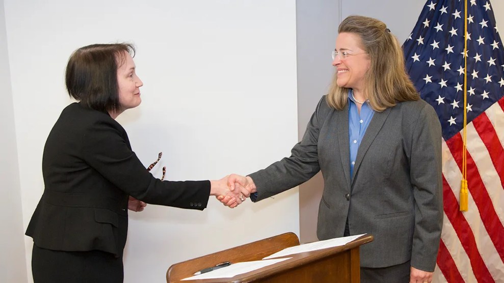 Annie Caputo and Kristine Svinicki shake hands over a wooden podium with an American Flag in the background.