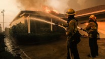 Two people in firefighting gear face away from the camera, spraying from hoses onto the roof of a house, which is on fire. Smoke billows above the home. The sky is gray.