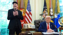 Donald trump wears a suit and sits behind a dark wood desk expression-less. Elon Musk stands next to the desk, smiling and clapping.