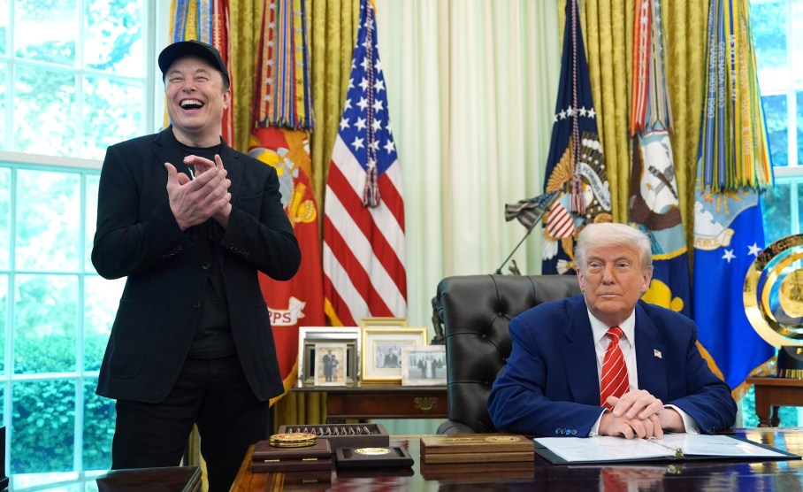 Donald trump wears a suit and sits behind a dark wood desk expression-less. Elon Musk stands next to the desk, smiling and clapping.