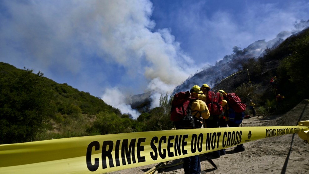 Close up shot of yellow tape stretched with the words: "CRIME SCENE DO NOT CROSS." Behind this barrier are three people dressed in yellow jackets and helmets with red backpacks. They are looking out at smoke billowing from the place where two green mountains meet against a blue sky.
