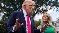 President Donald Trump in a suit and red tie gestures with his hands while speaking outside under a leafy tree. White House press secretary Karoline Leavitt stands beside him, slightly out of focus, looking at him.