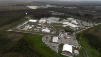 Aerial shot of white industrial buildings and parking lots surrounded by green grass and trees.
