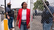 A woman in jeans and a red suit jacket exits a parking lot through a chain link fence, while flanked by two plainclothes masked federal agents.