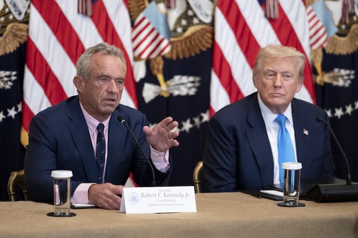 RFK Jr and Trump sitting at a table with American flags behind them and microphones in front of them. RFK Jr is speaking.