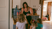 A young woman stands in a kitchen in front of three girls of descending height. The woman is facing the camera, looking down as she holds a blue ice pop and a pair of scissors. The children are seen from behind, looking up at the woman with rapt attention.