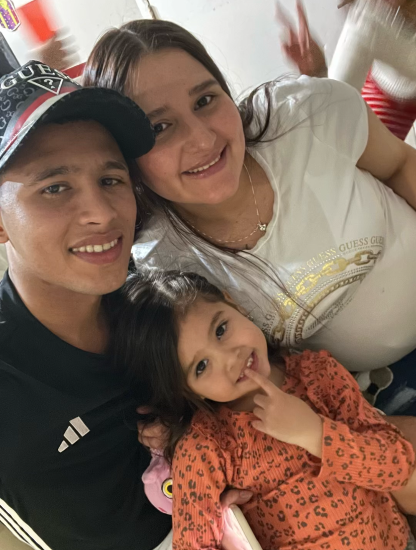 A photo of a man, woman and young girl posing closely together with smiles on their faces. In the background someone holds up the peace sign, using two fingers, in the upper right corner of the photo.
