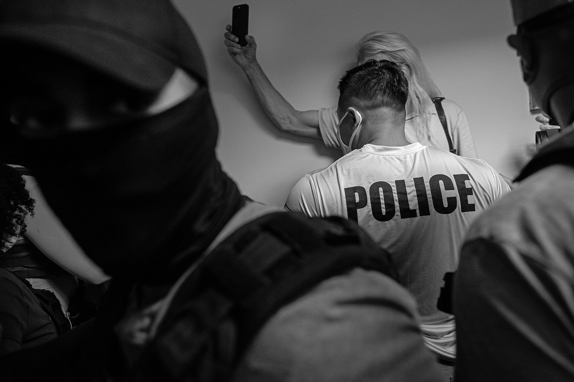 Federal agents face a woman holding a phone, against a wall.