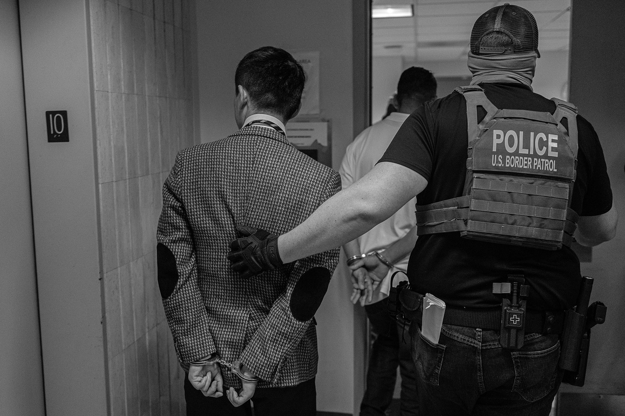 US Border Patrol agent leads a handcuffed man in a suit jacket through a door.