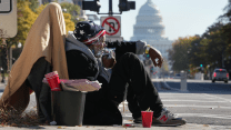 A man sits on a street corner amid a pile of his belongings on a sunny day. Smoke from a cigarette held to his mouth wafts past his face. In the background are trees lining a street and the dome of the US Capitol.