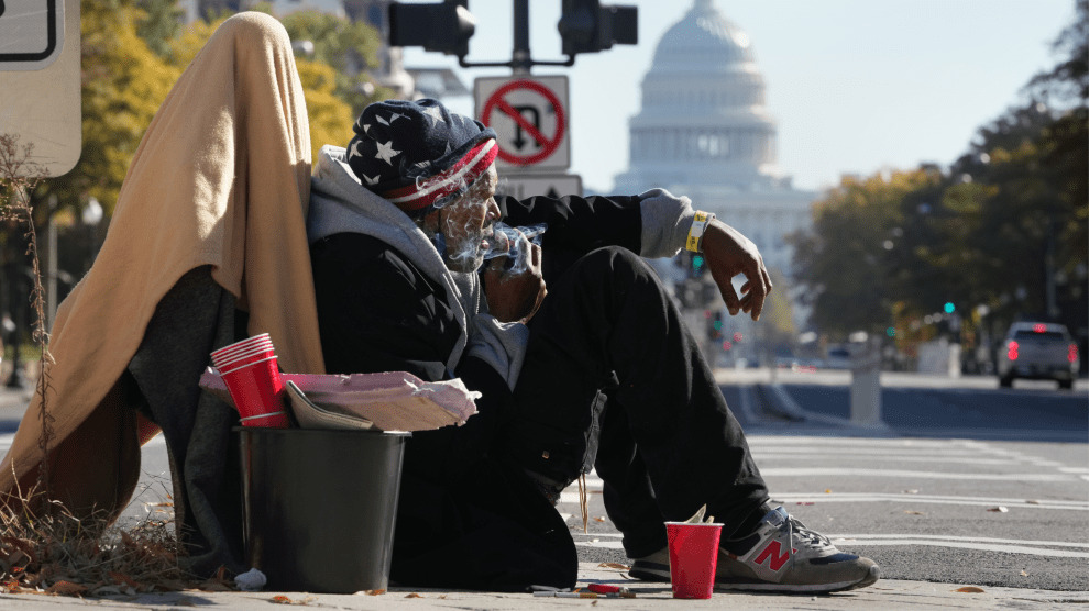 A man sits on a street corner amid a pile of his belongings on a sunny day. Smoke from a cigarette held to his mouth wafts past his face. In the background are trees lining a street and the dome of the US Capitol.