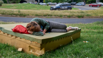 A woman in a green long-sleeve sweatshirt, gray pants and sneakers lays curled up on a foam mattress on a patch of unkept green grass. Her head rests on her right arm, a red bag lays above her head on the mattress.