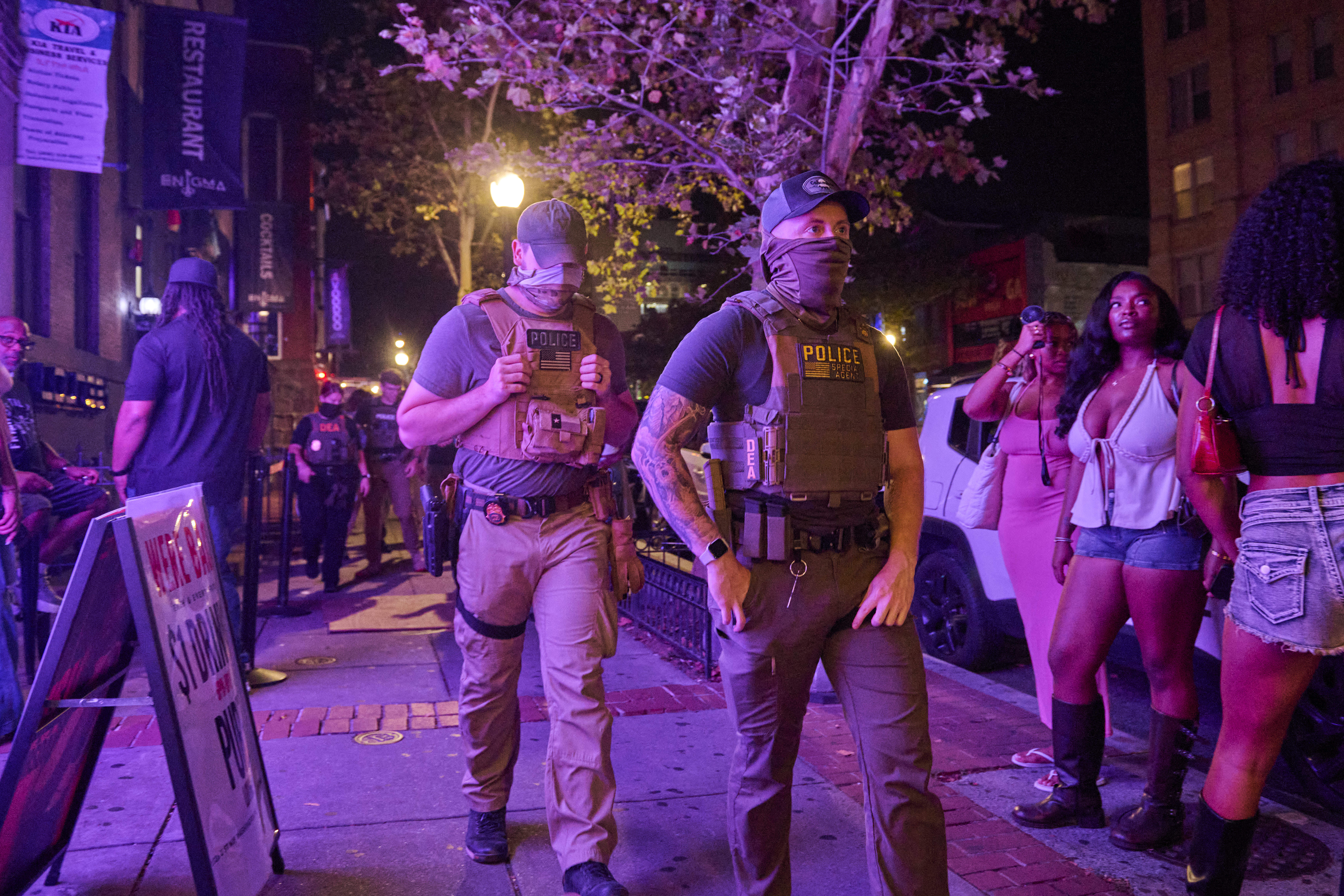 People dressed for a night out stand back from masked officers walking down the sidewalk.