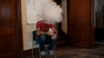 A man with tattoos on both arms in a red T-shirt, blue jeans, and sneakers sits in a chair in a room. The man’s face is obscured by a puffy cloud of smoke.