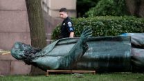 A uniformed police officer stands over a toppled copper statue of a bearded man.