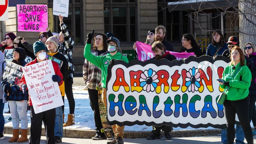 A group of protesters standing with a large rainbow sign that says Abortion is Healthcare, and smaller signs including one that says Abortions Save Lives