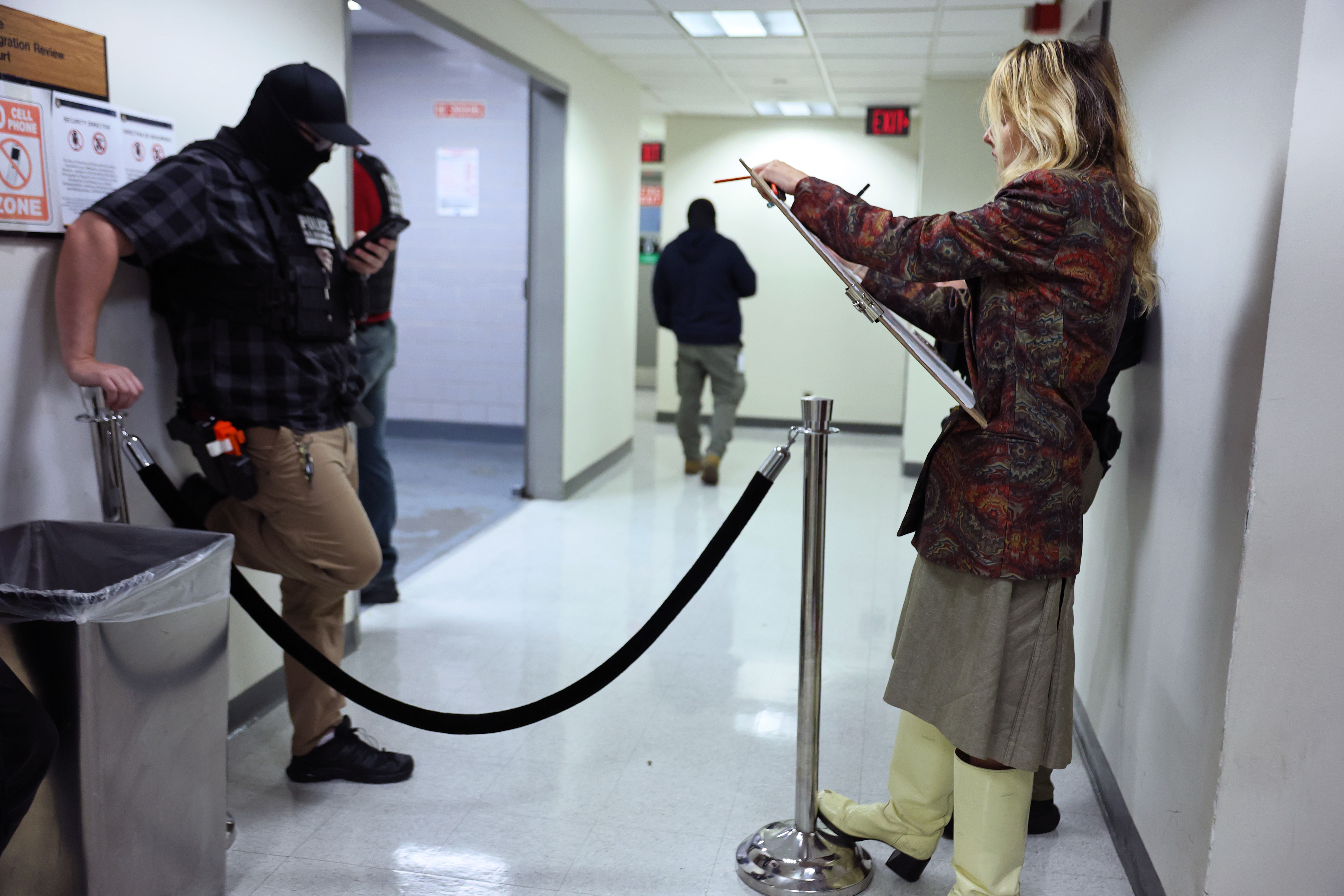 A woman with her sketch board leans against a wall in a hallway and draws while a masked law enforcement officer stands near her looking at his phone.