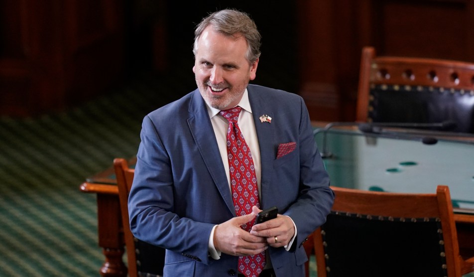 A Caucasian man in a blue suit and red tie stands in front of a heavy wood table, clasping his hands together, grinning.