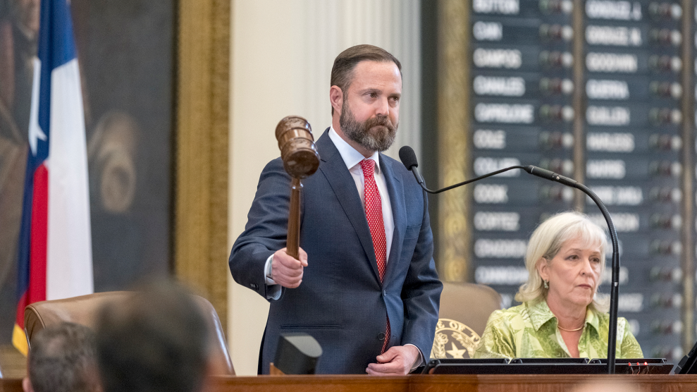 A Caucasian man, with a beard in a blue suit and a red tie, holds a gavel, as he stands on a podium in front the Texas flag. A Caucasian woman with blonde hair, wearing a lime-green shirt, is seated beside him.